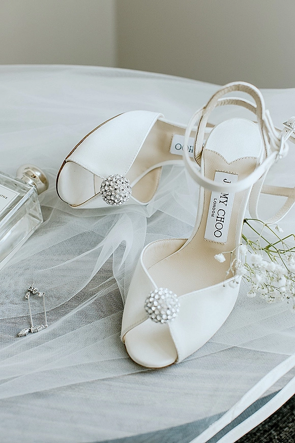 Bridal shoes and white wedding heels with crystal embellishments beside a tulle veil, perfume bottle, bracelet, and baby’s breath on a chair