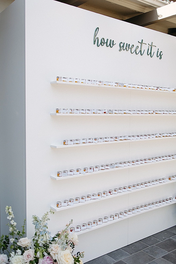 Wedding favor display with small favor jars and labeled stickers on shelving, accented by a floral arrangement and script wall sign on a white wall