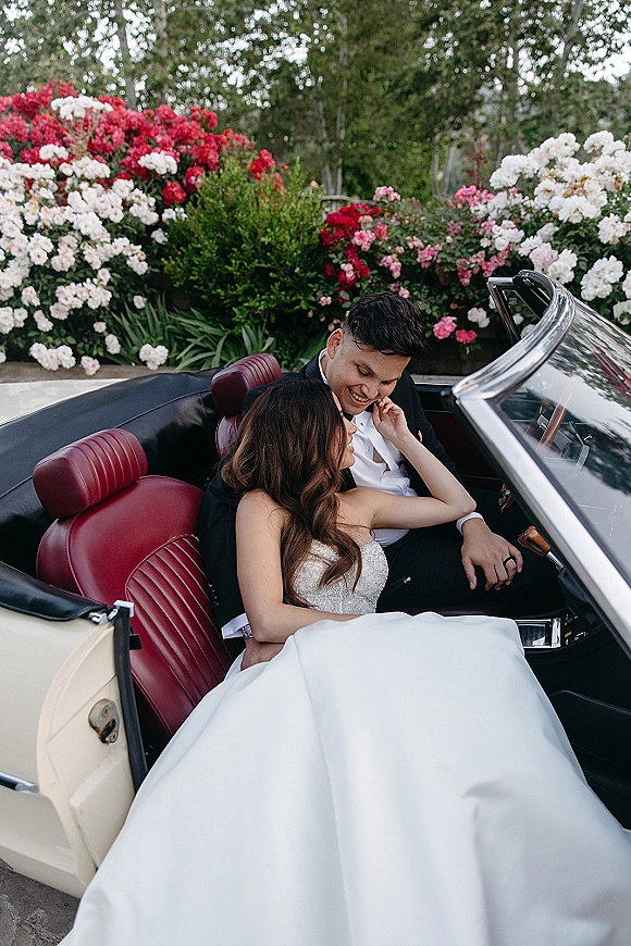 Couple portrait in a wedding getaway car, bride in strapless dress touching groom’s face amid red leather seats and garden greenery
