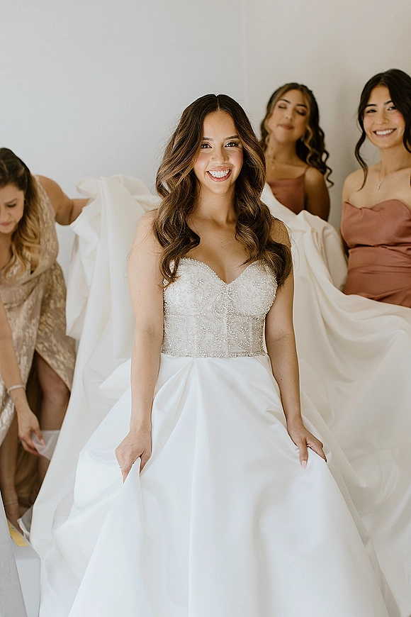 Bride getting ready in a bridal gown reveal, smiling as bridesmaids fluff her strapless beaded bodice dress in an indoor room with a white wall