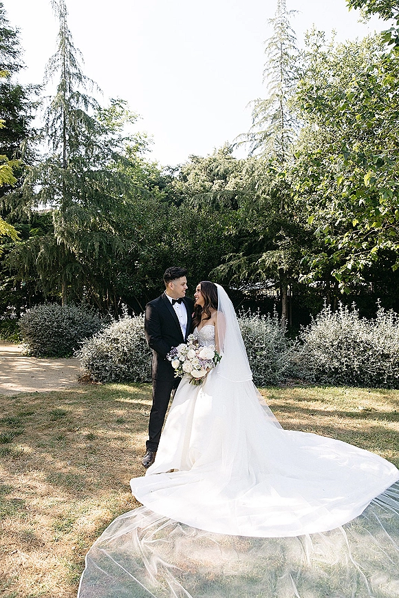 Couple portrait of bride and groom in a sunlit garden, she wears a cathedral veil and holds a white and lavender bouquet as they gaze at each other