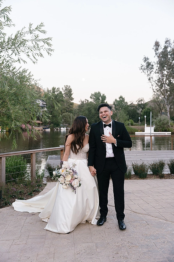 Couple portrait of bride and groom laughing, holding hands on a wooden dock by a lake, bride holding a bouquet in golden light
