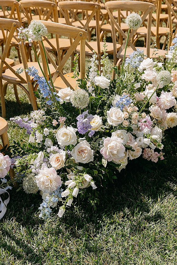 Ceremony aisle florals border a wedding aisle flower meadow of white, blush, and light blue blooms beside cross back chairs on a lawn