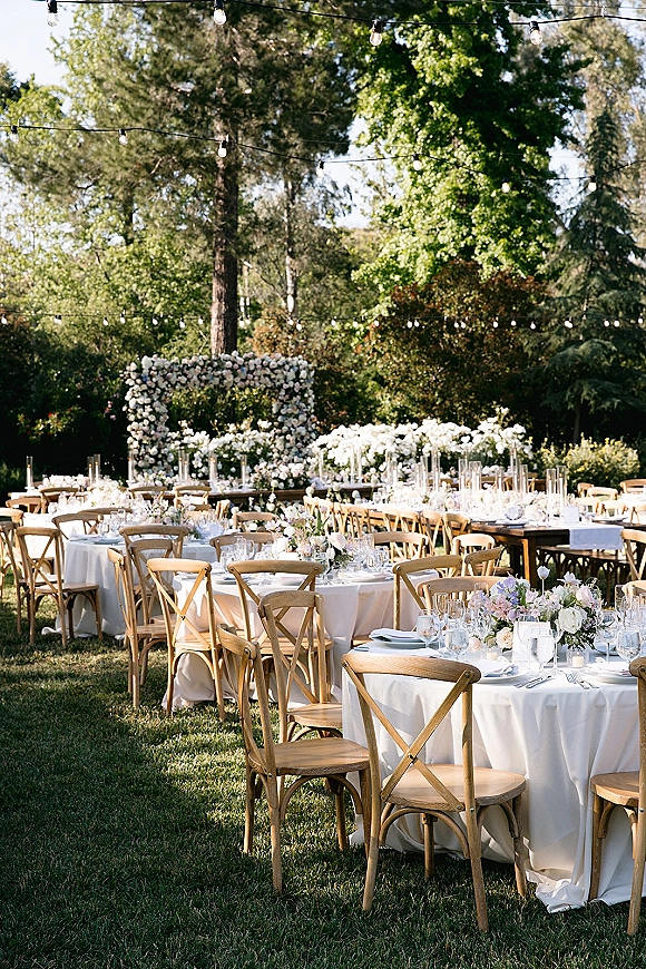 Outdoor wedding reception with garden wedding reception tables set in white linens, crossback chairs, floral arch, and string lights on a sunny lawn
