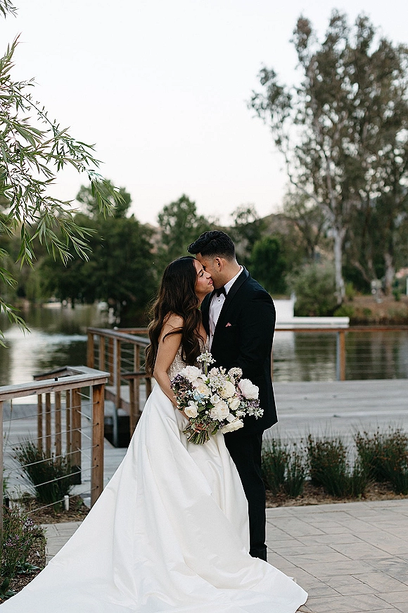 Wedding kiss portrait of bride and groom kissing on a lakeside dock, bride holding bouquet with veil and groom in black tuxedo