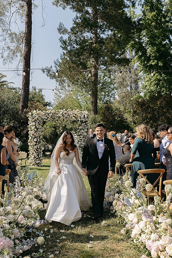 Wedding recessional as bride and groom walk the aisle beneath a floral arch, cathedral veil and tux, with string lights over garden guests