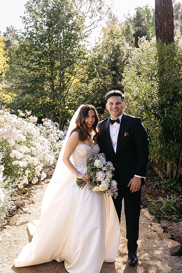 Couple portrait of bride and groom smiling on a sunlit garden path, bride in veil holding a bouquet beside groom in tuxedo