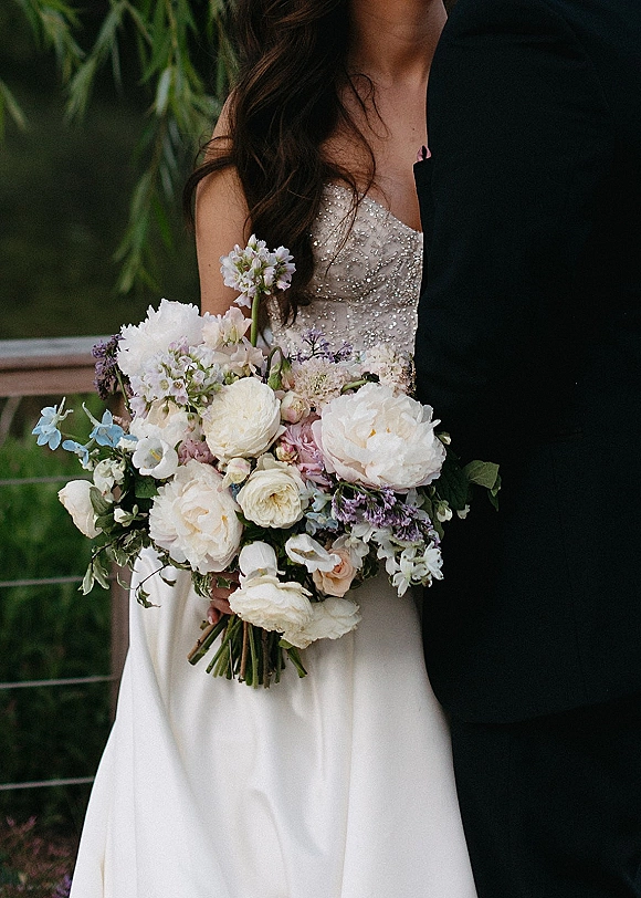 Bridal bouquet of white peonies and blush roses with lavender accents and greenery, held by a bride in a beaded strapless dress outdoors