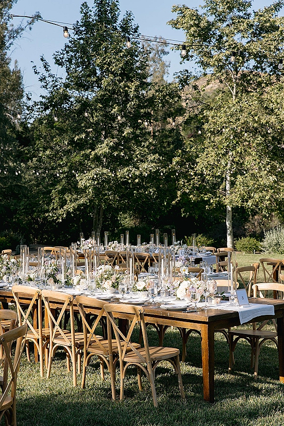 Reception tablescape for an outdoor wedding reception on wood farm tables with white runner, florals, candles, and string lights on lawn