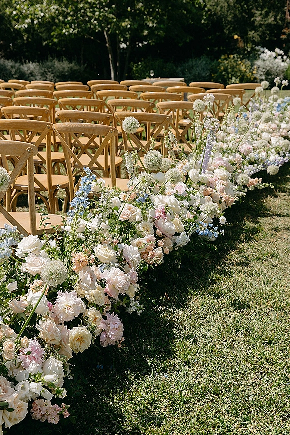 Ceremony aisle decor with outdoor ceremony aisle flowers in a low rose, peony, and blue delphinium arrangement beside wood chairs on a lawn
