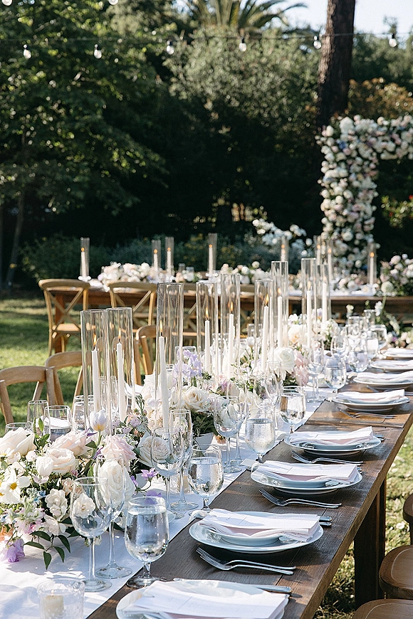 Reception tablescape with an outdoor wedding tablescape feel, long wood farm table set with white runner, rose garland, candles, string lights outdoors