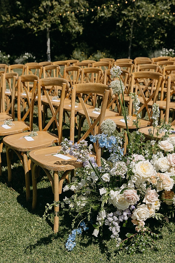 Ceremony seating with wood cross back chairs, reserved seat cards, and pastel aisle flowers on a garden lawn under string lights
