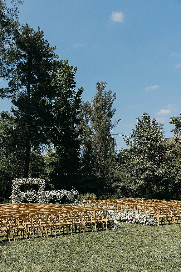 Ceremony setup for an outdoor wedding ceremony with wood crossback chairs lining an aisle of white flowers beneath a floral arch on a lawn under blue sky
