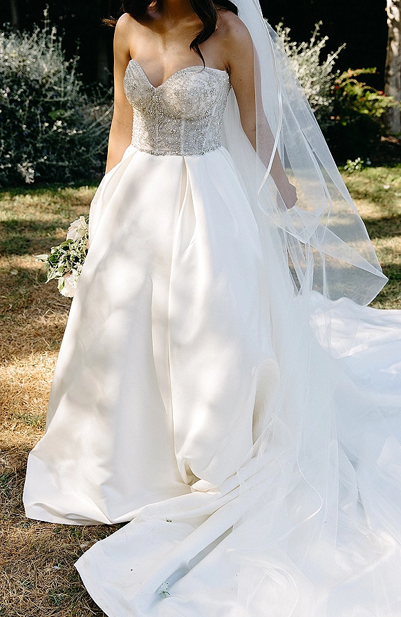 Wedding dress with strapless wedding dress beaded bodice, full skirt and long veil, held with bouquet on a sunlit garden lawn