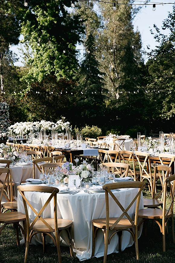 Outdoor wedding reception with round tables in white linens, floral centerpieces and votive candles under string lights on a garden lawn