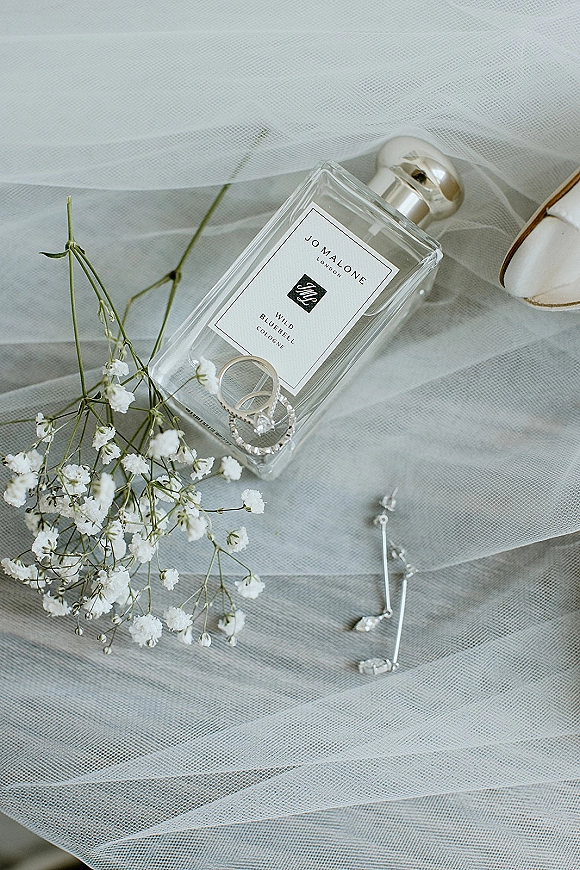 Wedding flat lay of wedding rings, perfume bottle, drop earrings and bridal shoes on a light fabric surface with baby’s breath accents
