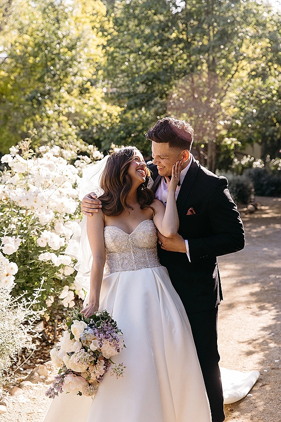 Couple portrait of bride and groom embracing and laughing, her long veil and bouquet glowing in golden hour light on a garden path.
