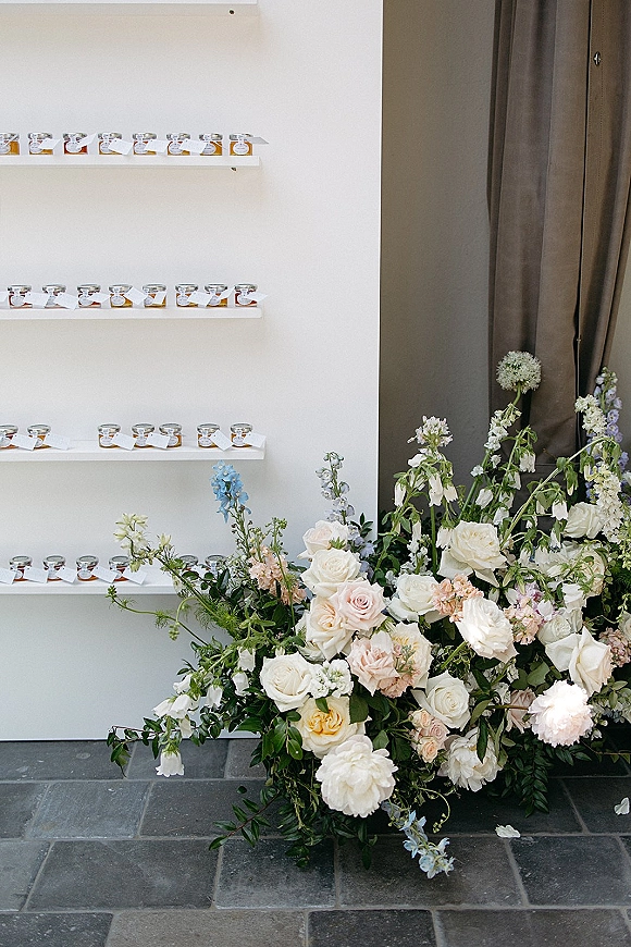 Wedding favor display with jar wedding favors and personalized tags on floating shelves beside pastel rose and peony arrangement against a white wall