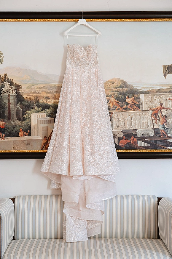 Wedding dress with lace overlay and strapless sweetheart bodice hangs on a hanger, long train falling beside framed painting and striped sofa