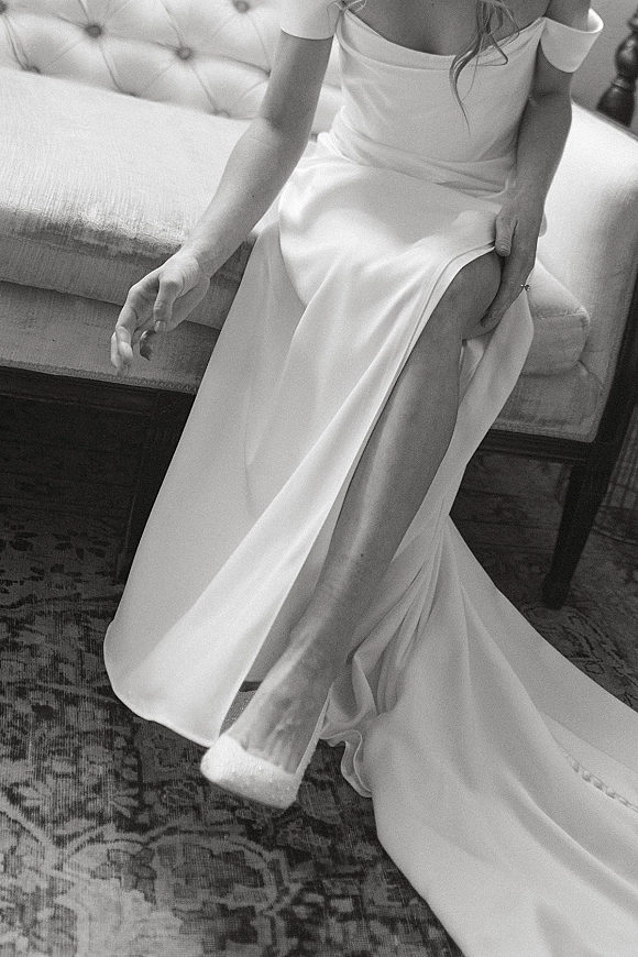 Wedding dress with off the shoulder neckline as the bride slips on pointed toe shoes, ring visible beside a tufted sofa