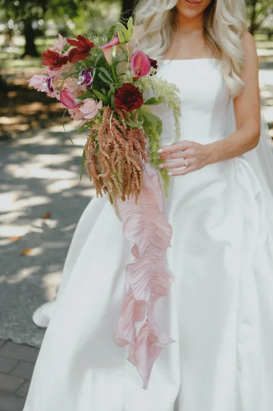 Bridal bouquet cascading bridal bouquet with pink calla lilies, orchids and amaranthus, held against a white gown in dappled sunlight