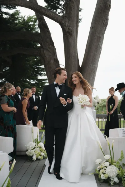 Ceremony recessional with a just-married aisle walk, the bride holding calla lilies as newlyweds smile on a tree-lined wooden deck among guests.