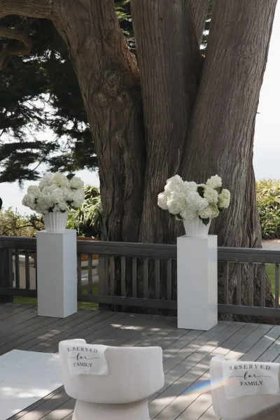 Ceremony decor with white pedestal florals frames the aisle runner on a wood deck under tall tree trunks facing a garden and ocean view.