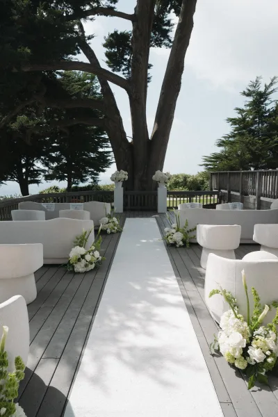 Ceremony setup with outdoor ceremony seating in lounge chairs on a wooden deck beneath a large tree, overlooking a calm ocean view.