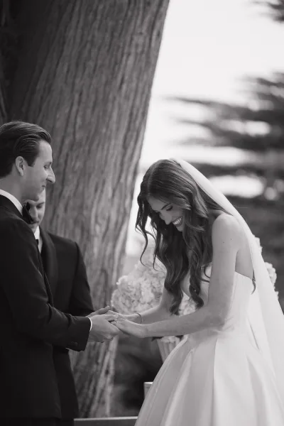 Ring exchange moment as the bride slips the wedding band onto the groom’s hand during a tender ceremony moment beneath leafy trees.