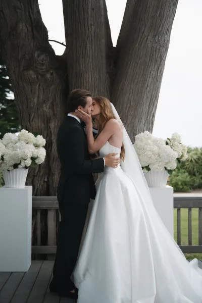 Wedding kiss portrait of a couple sharing their first kiss at the altar beneath a large tree, her long veil drifting above the wooden deck.