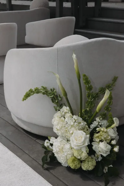 Wedding floral arrangement of white and green wedding flowers, featuring calla lilies, set by lounge chairs on a wood floor near a white runner.