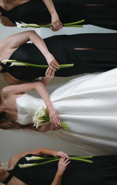 Bride and bridesmaids in black bridesmaid dresses stand arm in arm, each carrying a single calla lily for a sleek, modern monochrome vibe.