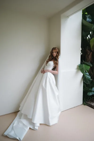 Bridal portrait of a bride in a strapless wedding dress, its long train fanned across a white architectural corner framed by tropical plants.