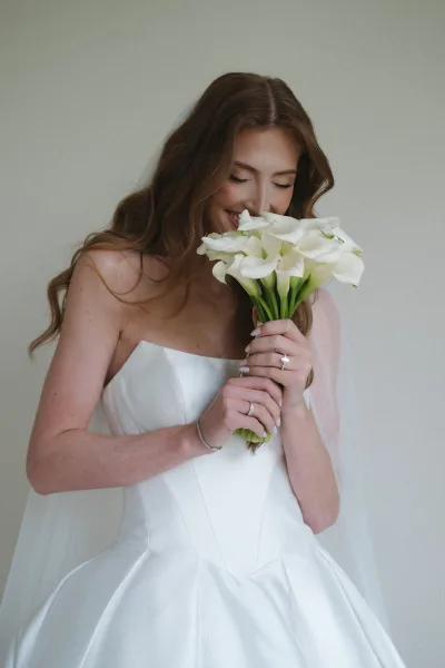 Bridal portrait of a bride with bouquet, cradling a white calla lily bouquet with a serene smile in a clean, minimalist bridal look.