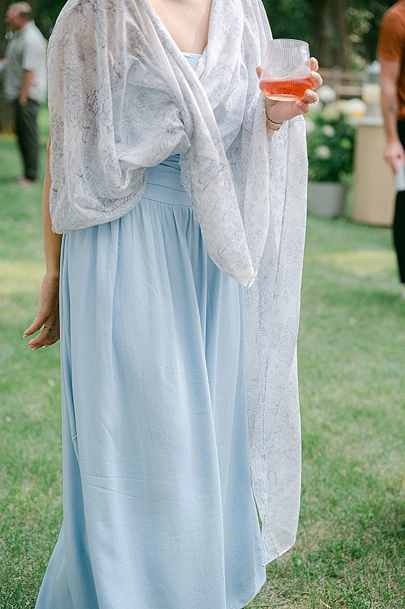 Wedding guest outfit in a blue wedding guest dress with sheer patterned shawl, holding a cocktail glass on an outdoor lawn with blurred guests and greenery