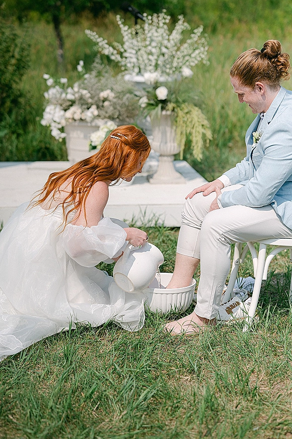 Wedding foot washing ceremony with bride kneeling to wash groom’s bare feet using a white pitcher and basin on an outdoor platform amid greenery