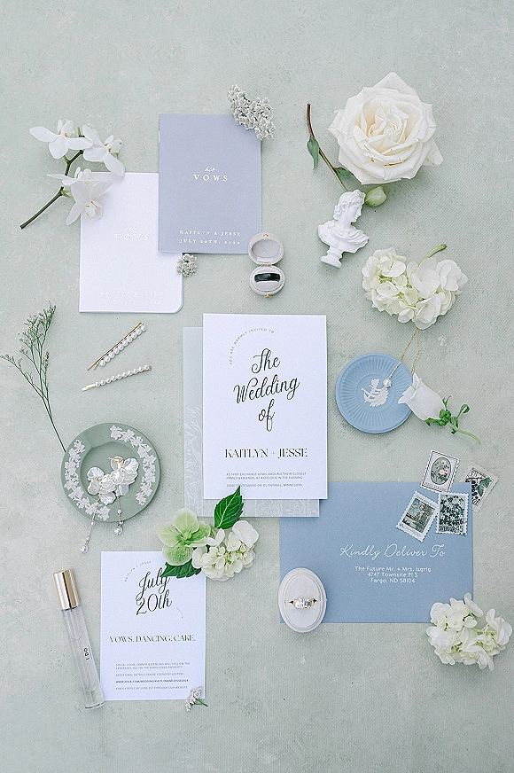 Wedding invitation suite with blue wedding invitations, calligraphy envelope, rings, wax seal, pearls and white blooms on sage linen backdrop