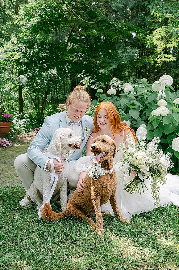 Couple portrait of bride and groom with dogs seated on a garden lawn, bride holding hydrangea bouquet, groom in light blue suit