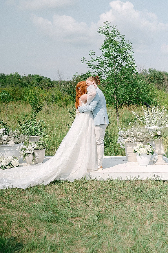 Wedding kiss as bride in strapless gown with long train and groom in suit embrace on a platform amid white florals, meadow sky behind