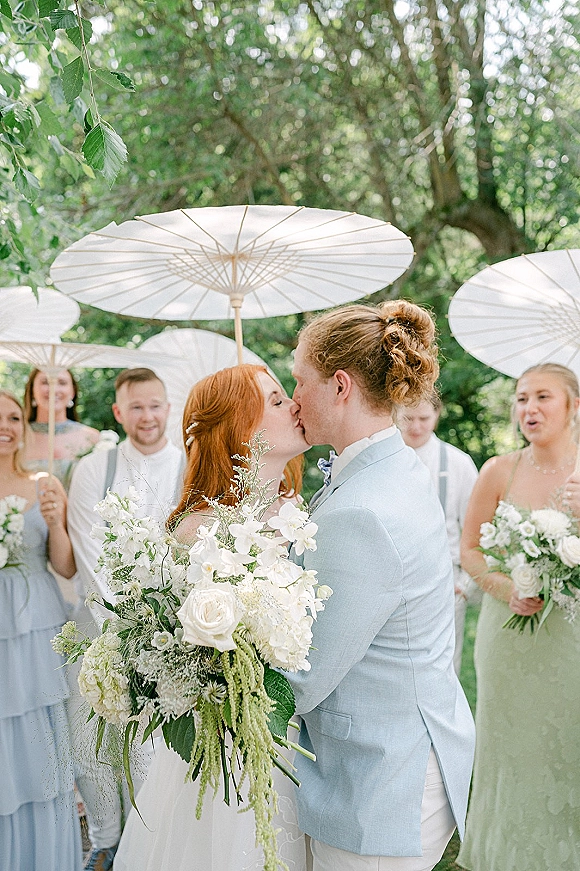 Wedding kiss as bride and groom embrace on a garden lawn, wedding party behind with white parasols and bouquets amid greenery