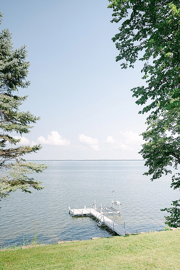 Lakefront wedding venue with a dock extending over calm lake water, framed by grassy shoreline, tree branches, and blue sky clouds