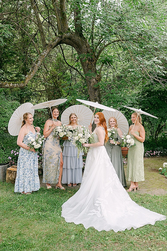 Bride with bridesmaids in a bridal party portrait, holding bouquets and white parasols on a lush garden lawn among trees and greenery