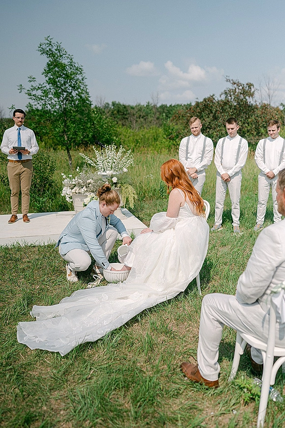 Wedding ceremony moment as groom kneels to wash the bride’s feet, her long train draped beside white chairs in a grassy field