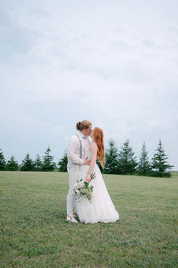 Wedding kiss portrait of bride and groom kissing, bride holding bouquet in a strapless dress, in a grassy field with evergreens under cloudy sky
