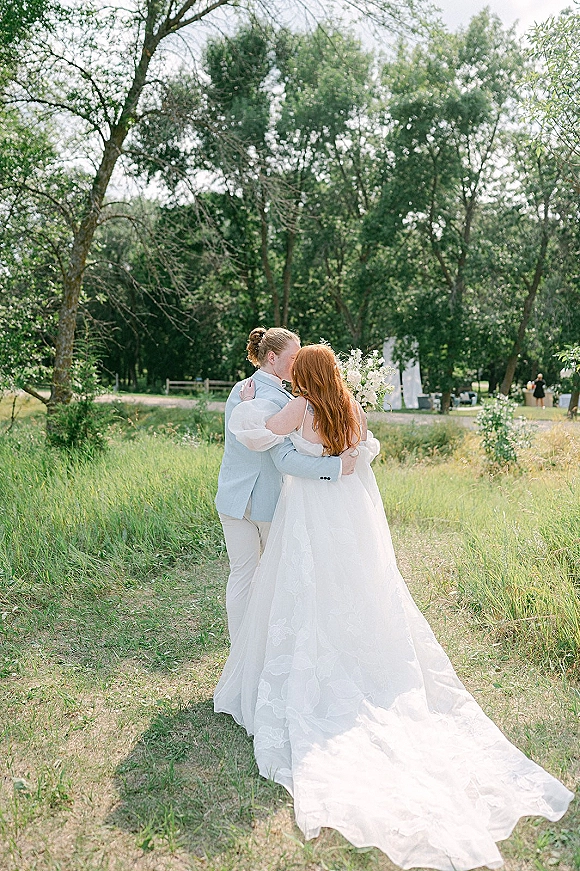 Wedding kiss portrait of bride and groom kissing on a dirt path, her long train flowing as she holds a bouquet in a meadow field