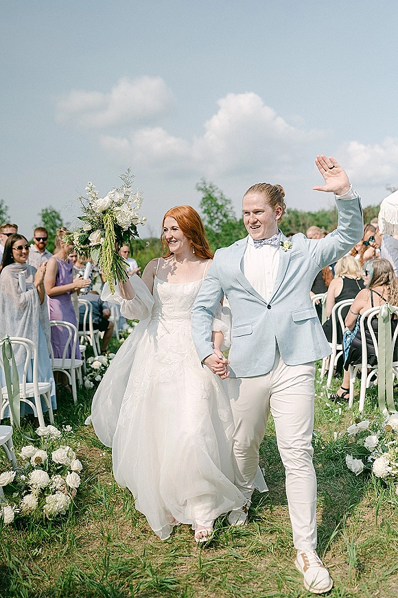 Wedding recessional as bride and groom walking aisle hand in hand, groom waving, bride holding bouquet under a cloudy sky outdoors