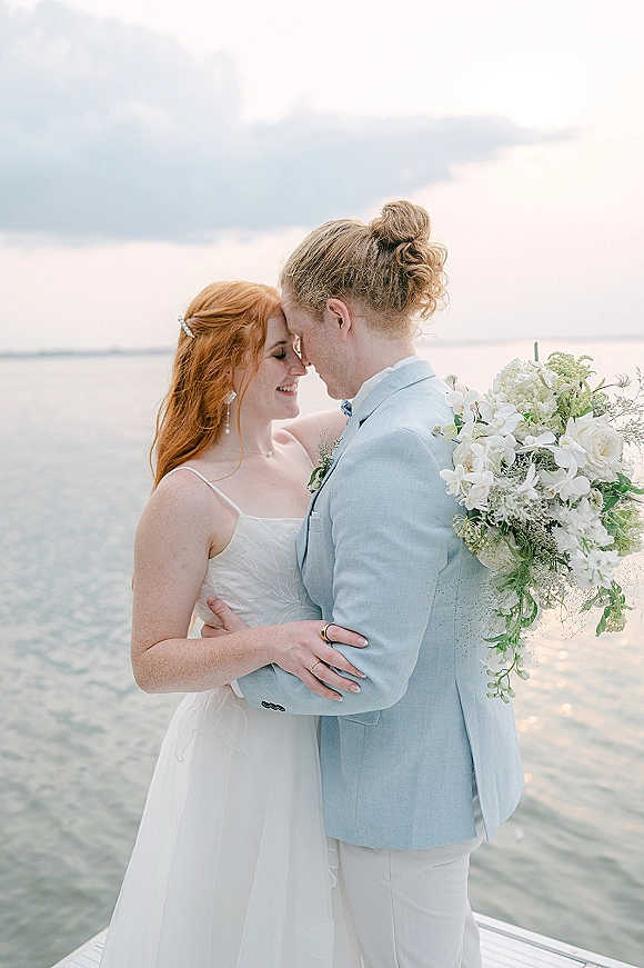 Couple portrait of bride and groom embrace with a forehead touch, her orchid bouquet and his light blue suit by a cloudy dock shoreline