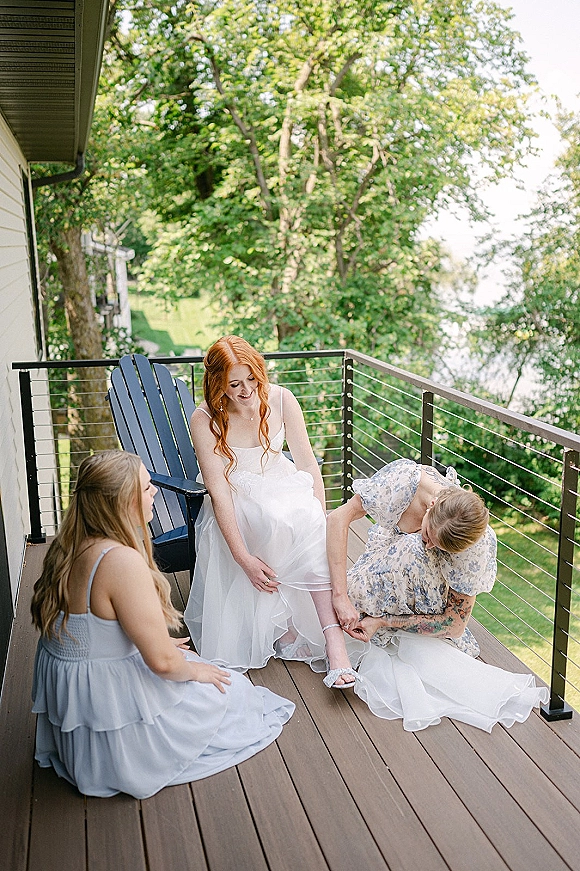 Bridal getting ready as bridesmaids help the bride put on white strappy heels beside an Adirondack chair on a lakeside deck