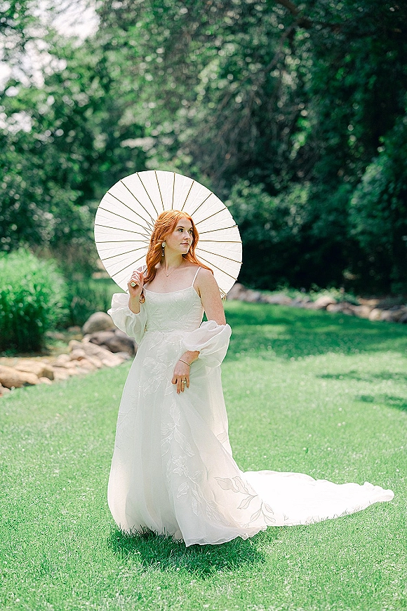 Bridal portrait of a bride with parasol, wearing a long-train wedding dress in a lush garden with trees and rocks, wearing drop earrings