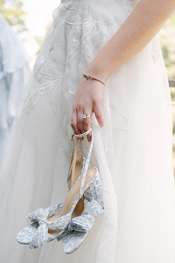 Bridal shoes in blue and white with bow slingback detail held over a white wedding dress, with ring and bracelet in soft greenery light